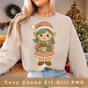 Puede incluir: Sudadera beige con una ilustración de una niña elfa sosteniendo una taza de cacao. La elfa lleva un gorro y un vestido rosa, una bufanda azul y tiene el pelo azul. El texto "Cozy Cocoa Elf Girl PNG" está en la parte inferior.