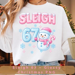 May include: White sweatshirt with a pink and blue design. The design includes the word "SLEIGH" in pink bubble letters, the number "67" in blue, and a cartoon snowman wearing a pink hat and scarf, holding a candy cane. Snowflakes and stars surround the design.