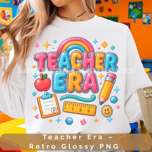 Puede incluir: Sudadera blanca con un diseño gráfico colorido de "Teacher Era". El diseño incluye un arcoíris, una manzana, un lápiz, una carpeta, una regla y una carita sonriente. El texto "Teacher Era - Retro Glossy PNG" está en la parte inferior.
