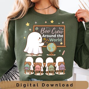 Puede incluir: Sudadera verde con un diseño de Halloween. El diseño presenta un fantasma de dibujos animados enseñando frente a una pizarra que dice "Boo Crew Around the World". La imagen incluye estrellas, un globo terráqueo y estudiantes con mochilas.
