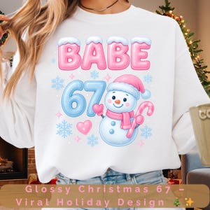Puede incluir: Sudadera blanca con un diseño festivo. El diseño presenta la palabra "BABE" en rosa y azul, el número "67" en azul y un muñeco de nieve con gorro y bufanda rosas. El texto "Glossy Christmas 67 - Viral Holiday Design" está en la parte inferior.