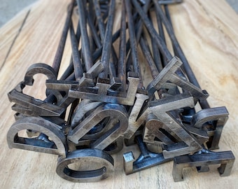 May include: A collection of dark gray metal branding irons, each featuring a unique letter or character at the end of a long, slender rod. The metal surfaces show a raw, textured finish with visible weld marks where the letters meet the rods.