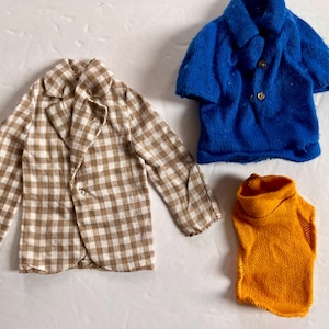 May include: Three pieces of doll clothing: a brown and white checkered blazer, a blue short-sleeved shirt, and an orange turtleneck sweater. The blazer has a two-button closure. The blue shirt has gold buttons.