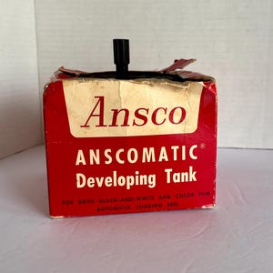 May include: A vintage red Anscomatic Developing Tank box with a black top. The box features the brand name "Ansco" and product details in white text. It is designed for both black-and-white and color film.