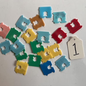 May include: A collection of twenty-three plastic bread clips in various colors including red, blue, green, yellow, and pink, scattered alongside a single white rectangular tag with the handwritten number 1 printed in black ink.