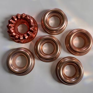 May include: Six metallic copper-colored baking molds arranged on a surface. One mold features a fluted, scalloped edge design, while the other five molds have a smooth, circular, ring-shaped structure with a central hole for baking cakes.