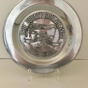 May include: A silver-colored plate with a raised rim. The center features a detailed emblem with the text "Erie County Bicentennial 1776-1976" and a map of the county. The words "Gift of Light" are also present.
