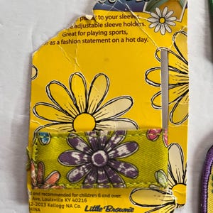 Peut inclure: Emballage en carton jaune pour attaches de manches avec des motifs de marguerites. L'attache de manche est verte avec une impression de marguerite violette. Le texte sur l'emballage indique "Ties" et "Great for playing sports, or as a fashion statement on a hot day."