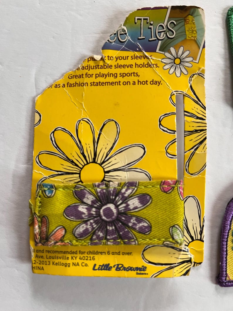 Peut inclure: Emballage en carton jaune pour attaches de manches avec des motifs de marguerites. L'attache de manche est verte avec une impression de marguerite violette. Le texte sur l'emballage indique "Ties" et "Great for playing sports, or as a fashion statement on a hot day."