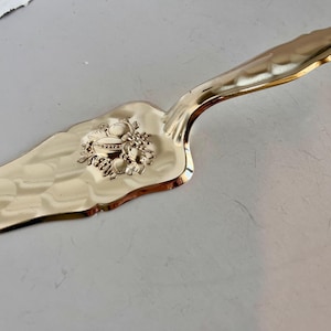 May include: A gold-colored cake server with a decorative handle and a detailed embossed design of fruit. The server has a scalloped edge and a curved handle for easy use. A kitchen utensil for serving cakes and pies.