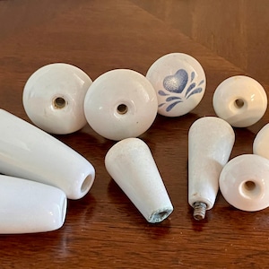 May include: A collection of white ceramic cabinet knobs and pulls. Some are round, others are cylindrical or cone-shaped. One knob has a blue floral design. The hardware is arranged on a wooden surface.