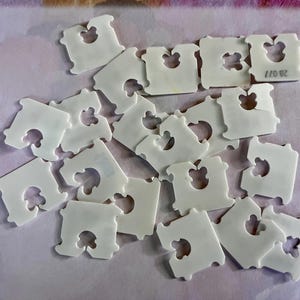May include: A collection of white plastic bread clips, each featuring a central cutout and a notched design for securing packaging. One clip displays the printed text 28 077. These small, rectangular fasteners are scattered in a loose arrangement.