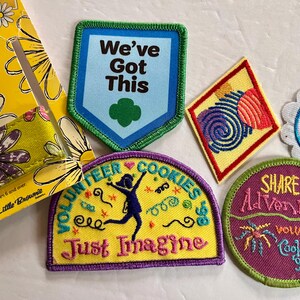 Peut inclure: Plusieurs &eacute;cussons brod&eacute;s color&eacute;s avec des motifs et des textes vari&eacute;s, dont "We've Got This", "Take Action" et "Share the Adventure". Comprend &eacute;galement un porte-manches jaune &agrave; motifs de marguerites.
