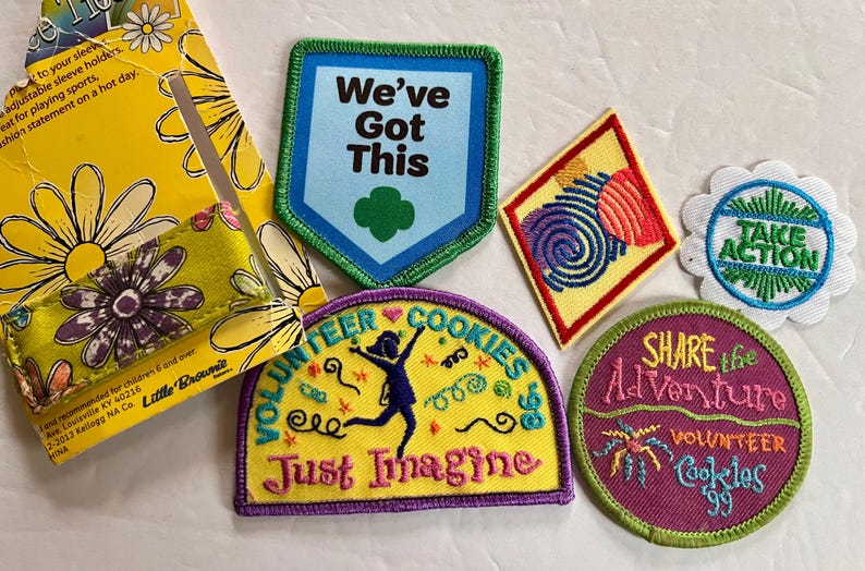 Peut inclure: Plusieurs &eacute;cussons brod&eacute;s color&eacute;s avec des motifs et des textes vari&eacute;s, dont "We've Got This", "Take Action" et "Share the Adventure". Comprend &eacute;galement un porte-manches jaune &agrave; motifs de marguerites.