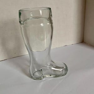 May include: Clear glass boot-shaped beer mug. The glass has a rounded top and a boot-like shape, with a foot and ankle. The glass is transparent, allowing the contents to be seen. The mug is designed for beer.