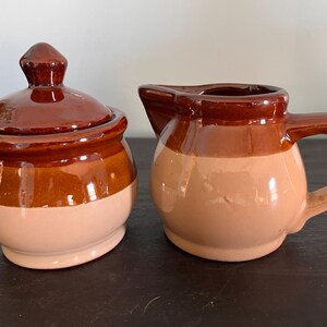 May include: A ceramic sugar bowl with a lid and a matching creamer pitcher. Both pieces feature a two-tone design with a glossy dark brown glaze on the upper sections and a smooth, light beige finish on the lower rounded bodies.