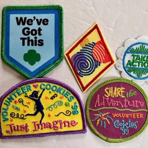 Peut inclure: Plusieurs &eacute;cussons brod&eacute;s avec des motifs et du texte vari&eacute;s. L'un d'eux porte l'inscription "We've Got This" avec un tr&egrave;fle vert. D'autres &eacute;cussons affichent les phrases "Take Action", "Volunteer Cookies" et "Share the Adventure".
