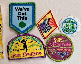May include: A collection of embroidered patches with various designs and text. One patch reads "We've Got This" with a green clover. Other patches say "Take Action", "Volunteer Cookies", and "Share the Adventure".