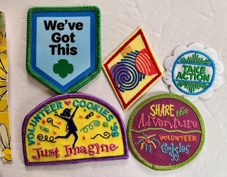 Peut inclure: Plusieurs &eacute;cussons brod&eacute;s avec des motifs et du texte vari&eacute;s. L'un d'eux porte l'inscription "We've Got This" avec un tr&egrave;fle vert. D'autres &eacute;cussons affichent les phrases "Take Action", "Volunteer Cookies" et "Share the Adventure".