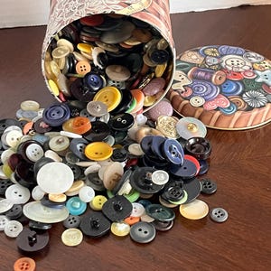 May include: A cylindrical metal tin with a decorative button-themed lid, tipped over to spill a large collection of assorted plastic and metal buttons in various colors, shapes, and sizes across the surface. The tin features a lace-like pattern.