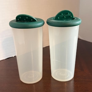 May include: Two clear plastic spice shakers with green lids. The lids have a flip-top design and are dark green. The shakers are cylindrical and sit on a dark brown wooden surface.
