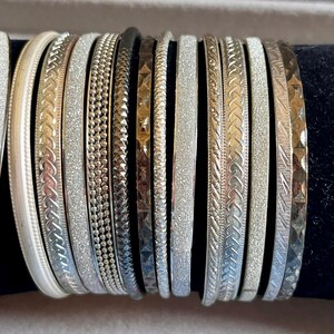 May include: A collection of thirteen thin metal bangles in various silver, gold, and dark metallic tones. The bracelets feature diverse textures including smooth surfaces, glittery finishes, beaded patterns, braided designs, and geometric engravings.