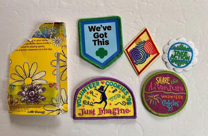 Peut inclure: Un ensemble d'&eacute;cussons et d'attaches de manches color&eacute;s. Les &eacute;cussons pr&eacute;sentent des textes tels que "We've Got This" et "Volunteer Cookies". Les attaches de manches sont jaunes avec des motifs de marguerites et du texte.