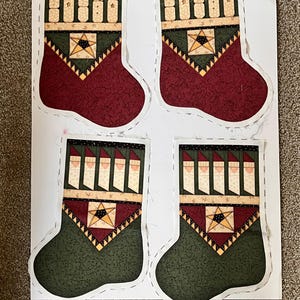 May include: Four fabric Christmas stocking panels printed on a single sheet. The top two feature maroon bodies with cream-colored snowman motifs at the cuff. The bottom two feature dark green bodies with cream-colored Santa Claus motifs at the cuff.