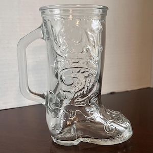 May include: Clear glass boot-shaped mug with a handle. The mug features embossed decorative patterns on the front and sides. The glass has a smooth, transparent appearance.