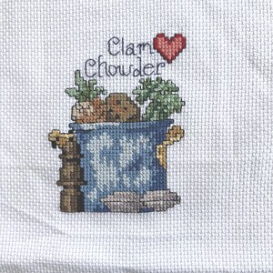 May include: A white fabric featuring a cross-stitch design of a blue pot filled with ingredients. The text "Clam Chowder" is stitched above the pot in black thread, accompanied by a small red heart. The design includes green herbs and brown items.