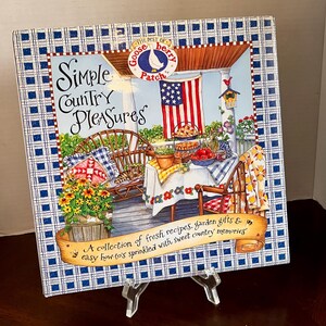 May include: A square cookbook titled "Simple Country Pleasures" with a blue and white checkered border. The cover features a country porch scene with a table set for a meal, an American flag, and the text "The Best of Gooseberry Patch."