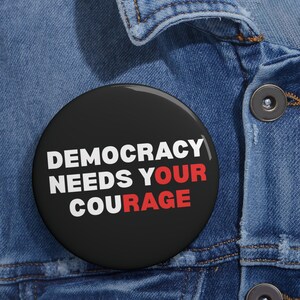 May include: A round, black button with the words "DEMOCRACY NEEDS YOUR COURAGE" in white and red lettering. The button is pinned to a blue denim jacket.