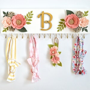 May include: A white wooden headband holder with gold hooks and floral embellishments. The holder features the letter "B" in gold lettering and is decorated with pink felt flowers and green felt leaves. Three headbands are hanging from the hooks, one yellow and white striped, one pink and white striped, and one white with a floral pattern.