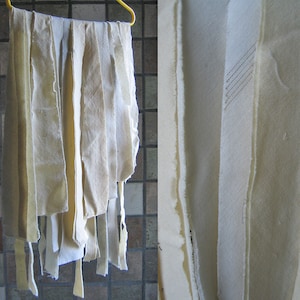 Puede incluir: Una colección de tiras de tela de color blanco roto colgadas de una percha amarilla. Las tiras de tela son de diferentes longitudes y anchos, y algunas tienen bordes deshilachados. La tela es de color natural sin blanquear.