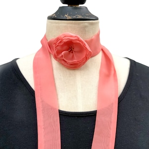 May include: A coral-colored scarf with a large, fabric flower accent. The flower is centered on the scarf and features multiple layers of petals with a cluster of small, brown beads in the center. The scarf is draped around a mannequin.