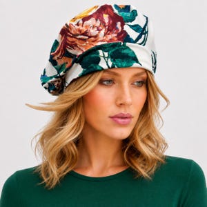 May include: A white beret featuring a large, vibrant floral pattern with deep red, orange, and yellow petals contrasted by dark green leaves and stems. The hat has a soft, rounded shape that sits comfortably on the head, showcasing a bold botanical print.
