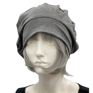 Op de afbeelding: Een grijze, losse beanie-muts van zachte stof. De hoed wordt getoond op een mannequinhoofd met kort, grijs haar. De beanie heeft een geplooide kroon en een brede, gevouwen rand, wat een casual en comfortabele stijl biedt.