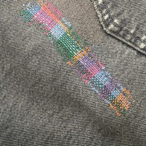 May include: Close-up of gray denim fabric with a colorful, embroidered design. The embroidery features a rectangular pattern with shades of pink, blue, green, orange, and purple. A denim pocket is visible in the upper right corner.