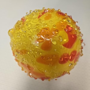 Puede incluir: Objeto decorativo de vidrio, de forma esférica, con base amarilla y detalles en naranja y rojo. La superficie está cubierta de pequeñas gotas transparentes. El objeto tiene una apariencia texturizada. Probablemente sea una pieza decorativa o de arte en vidrio.