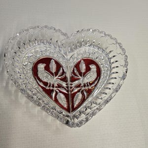 May include: A heart-shaped glass dish with a scalloped edge. The centre has a red panel with two white birds and leaf designs. The dish is likely for decorative use.