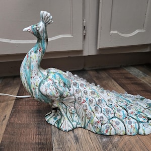 以下が含まれることがあります： 白を基調に青、緑、赤のペイントが施されたセラミック製の孔雀の置物。孔雀の尾には透明な丸い装飾が施されています。白い電気コードがベースから伸びています。