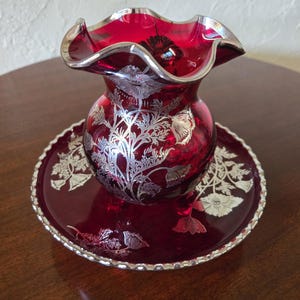 以下が含まれることがあります： ルビーレッドのガラス花瓶。縁はフリル状で、銀色の花のディテールが施されています。花瓶は、銀色の縁と花の模様が施されたお揃いのプレートの上に置かれています。家庭用の装飾品です。