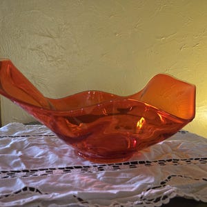 May include: An orange glass bowl with a wavy, organic shape. The bowl sits on a white lace tablecloth. The bowl's color is a vibrant, translucent orange, and it appears to be made of a single piece of glass.