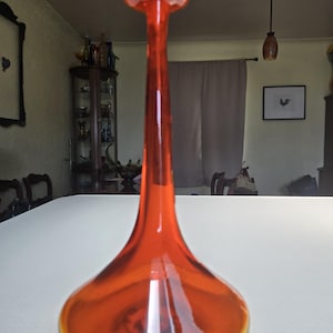 Pode incluir: Um vaso de vidro laranja vibrante com uma base larga e arredondada que se estreita num pescoço longo e fino, terminando num pequeno rebordo circular. O material translúcido capta a luz, realçando a sua silhueta suave e curva.