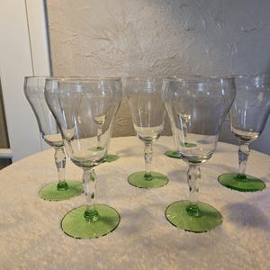 May include: Six clear wine glasses with green bases are arranged on a white surface. The glasses have a classic design with a flared bowl and a slender stem. The green bases add a pop of colour to the glassware.