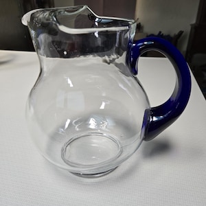 Puede incluir: Una jarra de vidrio transparente con un asa azul. La jarra tiene un cuerpo redondo y una boca ancha.