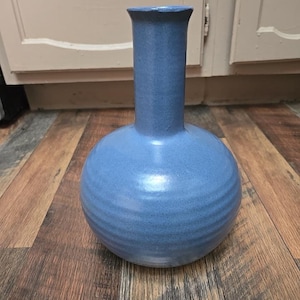 May include: A blue ceramic vase with a round base and a long, cylindrical neck. The vase has a matte finish and horizontal ridges around the base. The vase is sitting on a wooden surface.