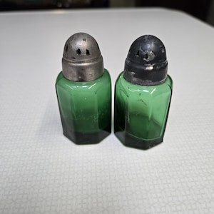 Puede incluir: Dos saleros y pimenteros de vidrio verde con tapas plateadas. Los saleros tienen forma hexagonal y están hechos de vidrio verde.