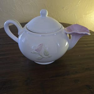 Op de afbeelding: Een witte keramische theepot met een bloemenontwerp. De tuit is gevormd als een roze bloem. De theepot heeft een afgeronde body, een gebogen handvat en een deksel met een kleine knop. Aan de zijkant is een roze en groen bloemenontwerp geschilderd.
