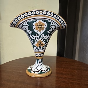 以下が含まれることがあります： 扇形の陶器製花瓶で、上部の縁が広がっており、円形の模様が施されています。本体は白地で、緑と黄色の花柄が描かれています。台座は円形で、黒いインクで手書きの署名が入っています。